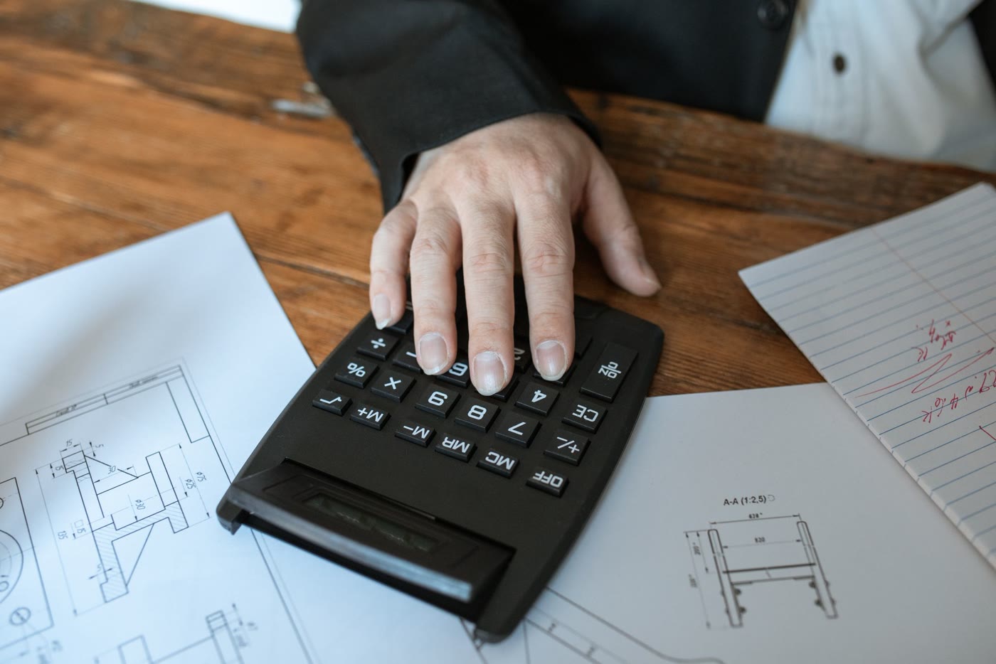 Mão sobre uma calculadora ao lado de plantas técnicas em uma mesa
